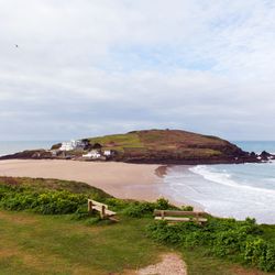 A scenic coastal view with a sandy beach, rolling green hills, and ocean waves. Two wooden benches sit on a grassy cliff overlooking the shoreline.