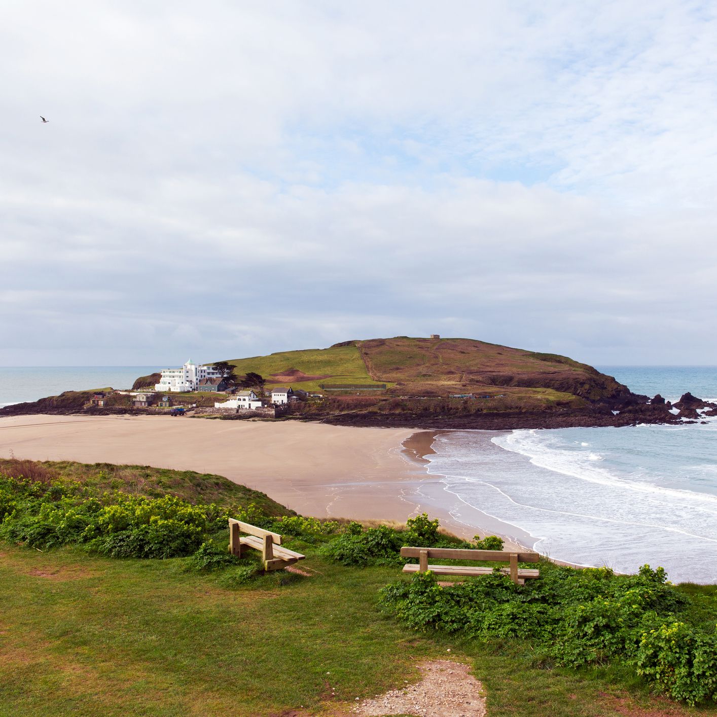 A scenic coastal view with a sandy beach, rolling green hills, and ocean waves. Two wooden benches sit on a grassy cliff overlooking the shoreline.