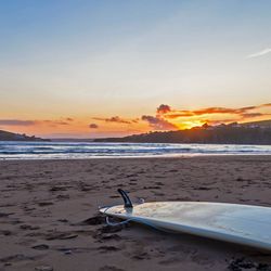 A surfboard rests on the sandy beach at sunset, with gentle ocean waves and a distant island in the background. The sky glows with warm orange and pink hues.