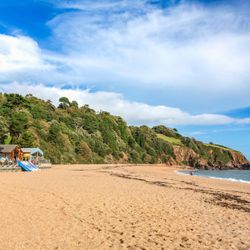 A sandy beach with gentle waves, backed by lush green hills and red cliffs. Small beach huts and kayaks sit near the shore under a bright blue sky with wispy clouds.
