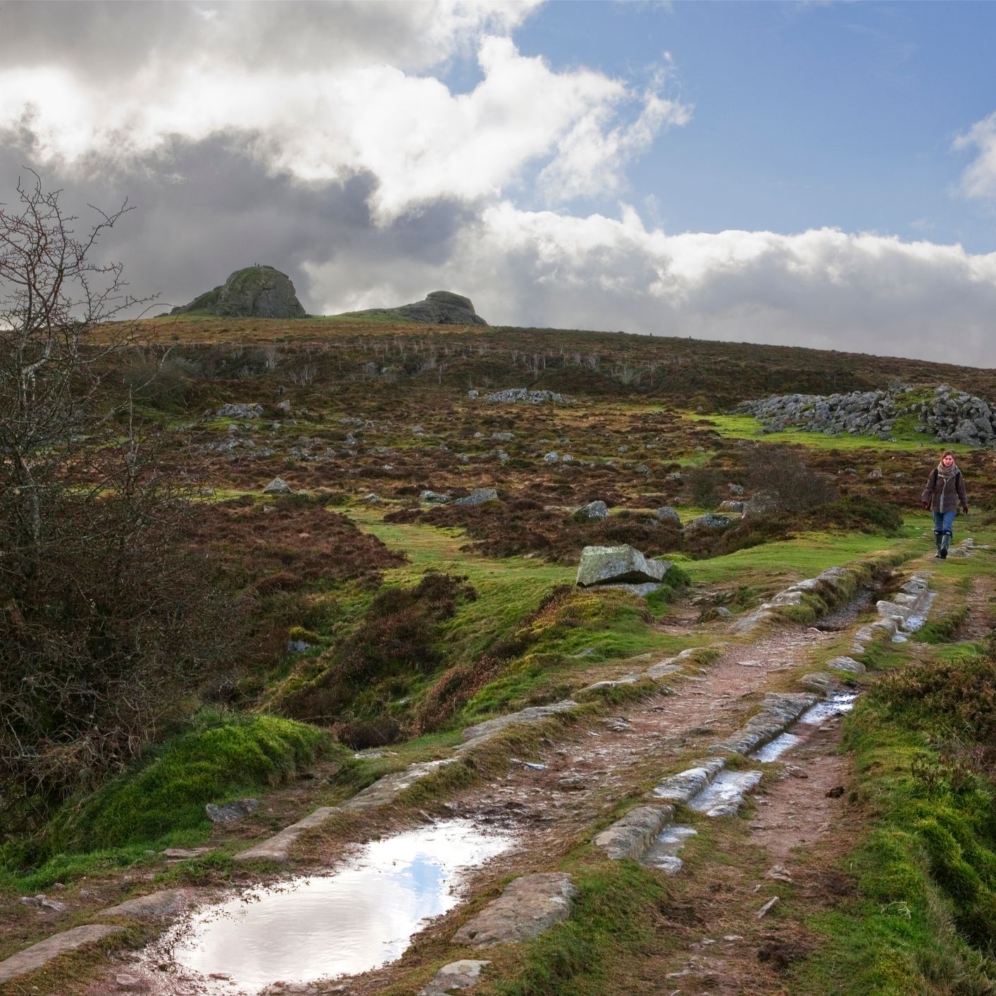 A lone hiker walks along a rugged, puddle-filled trail through a windswept moorland, with rocky hills in the distance under a cloudy sky.