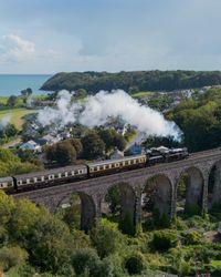 A steam train crosses a stone viaduct, releasing white smoke into the sky. Below, a village with green fields stretches toward the blue sea in the background.