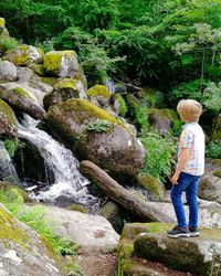 Two children stand on moss-covered rocks, admiring a small cascading waterfall flowing through a lush green forest, surrounded by large boulders and dense foliage.