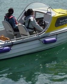 A small motorboat with a yellow canopy and purple fenders moves through calm turquoise waters. Two people wearing life jackets sit at the helm, steering the boat.