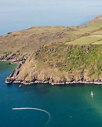 An aerial view of a coastal landscape with cliffs, green fields, and a sandy beach. The deep blue sea surrounds the peninsula, with a boat leaving a curved wake.