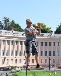 A man stands among a detailed miniature model of a grand, curved building, adjusting a tiny figure. The scene includes small lampposts, cars, and figurines.