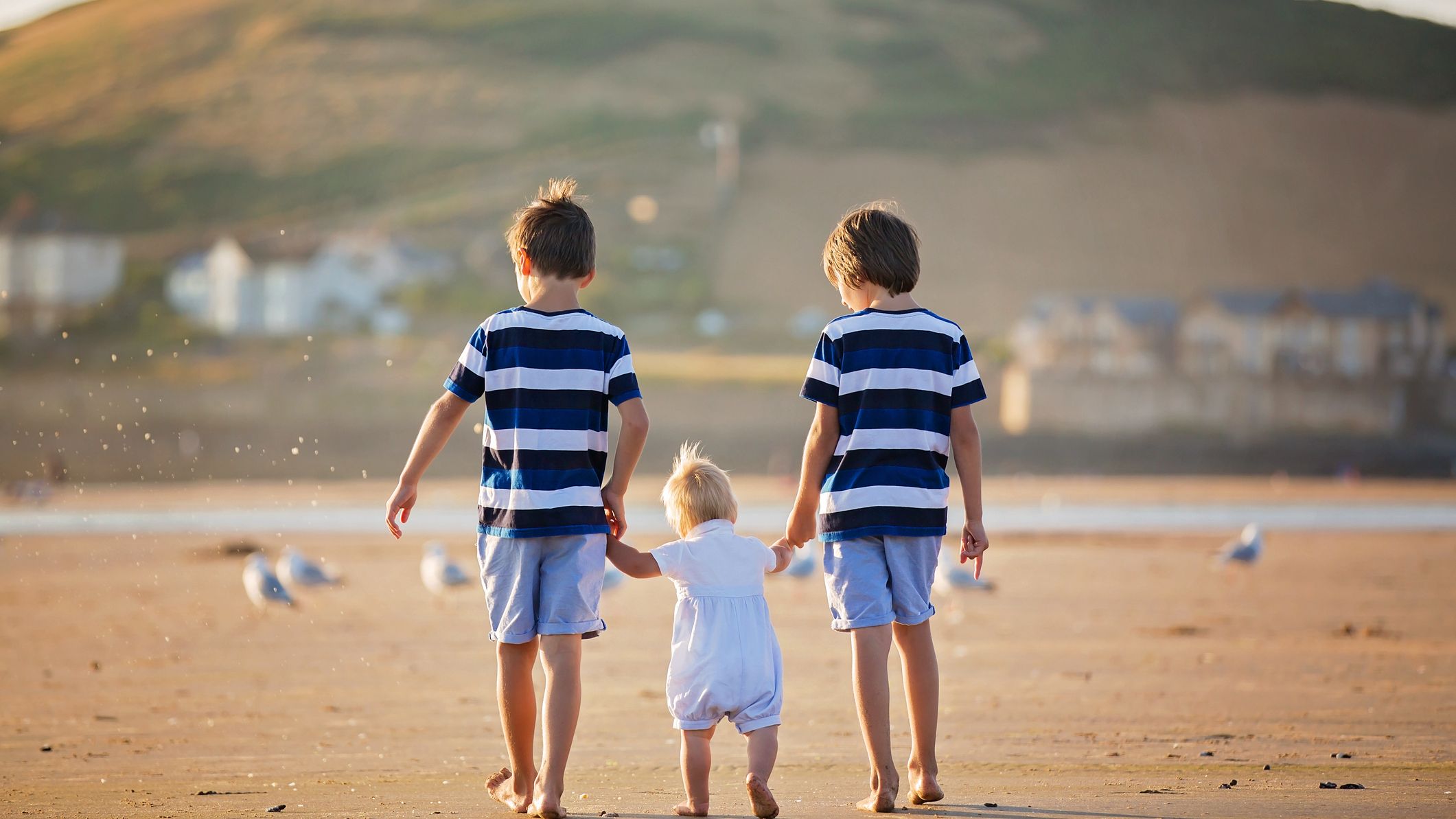 Three young siblings walk barefoot on a sandy beach, with the older two holding the toddler’s hands. The golden light and rolling hills create a peaceful backdrop.