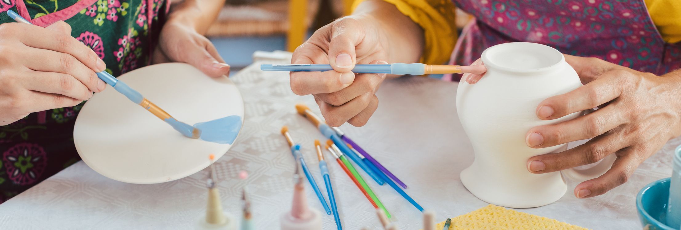 Two people paint ceramic pottery, one decorating a plate with blue paint and the other adding pink to a vase. Brushes, paint bottles, and water cups cover the table.