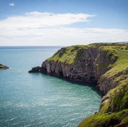 Rocky cliffs covered in green grass overlooking a calm turquoise sea, with a small island nearby under a partly cloudy blue sky.