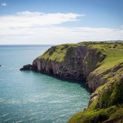 Rocky cliffs covered in green grass overlooking a calm turquoise sea, with a small island nearby under a partly cloudy blue sky.