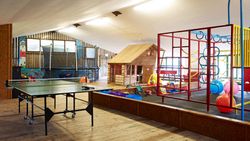 Indoor play area with a wooden playhouse, climbing frame, trampoline, ping pong table, and various toys on a wooden floor under a sloped ceiling.