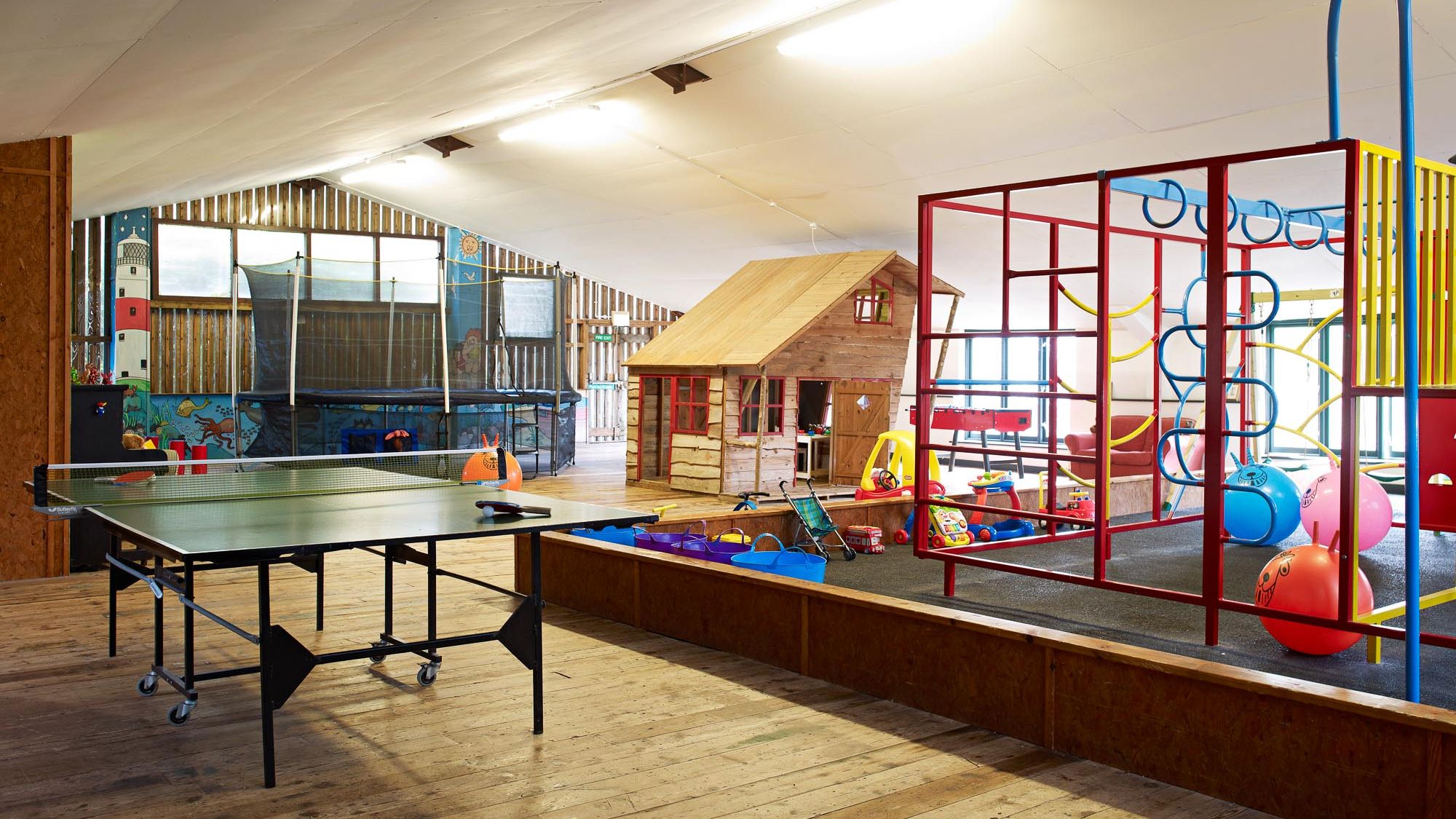 Indoor play area with a wooden playhouse, climbing frame, trampoline, ping pong table, and various toys on a wooden floor under a sloped ceiling.