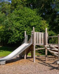 A wooden playground structure with a metal slide, climbing logs, and a small platform, set on wood chips and surrounded by lush green trees in an outdoor park.