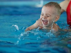 A joyful baby splashes in a swimming pool, laughing with eyes closed, while being supported by an adult wearing a red swimsuit in the background.