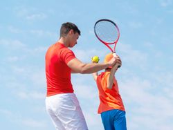 A tennis coach in a red shirt guides a young child in an orange outfit on how to swing a racket, against a bright blue sky with scattered clouds.