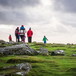 A family, including children, hike across a rocky, grassy moorland under a cloudy sky, dressed in bright winter gear.