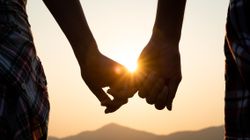 Close-up of a couple holding hands in front of a sunset, light flaring between their fingers with distant hills in the background.