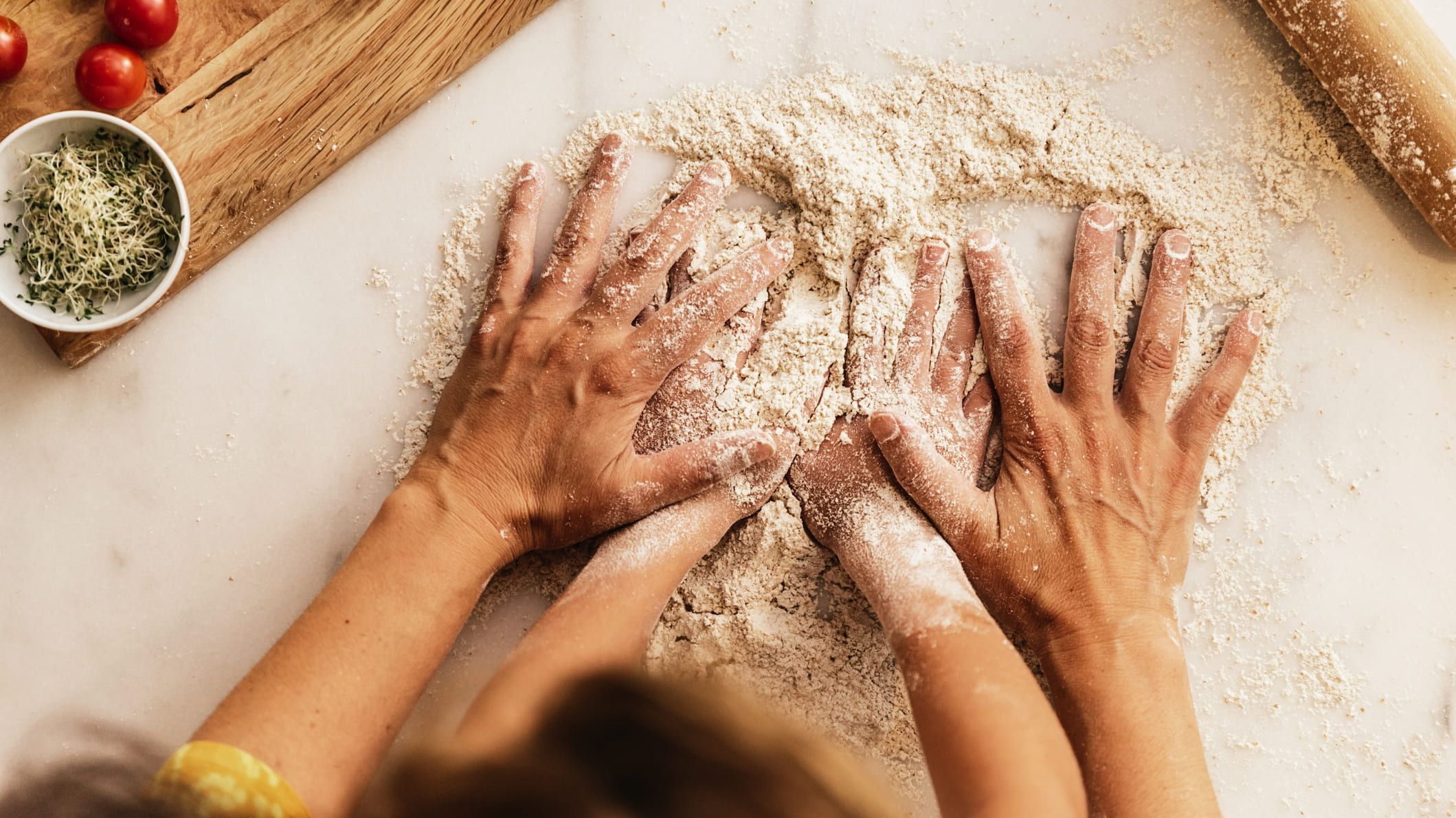 Adult and child hands working together to knead flour on a counter, surrounded by fresh ingredients like tomatoes, herbs, and a rolling pin.