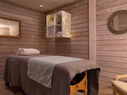 A cozy spa room with wooden walls, a massage table covered in soft linens, neatly rolled towels, warm lighting, a wicker mirror, and a shelf decorated with fairy lights.