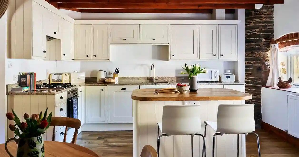 Spacious kitchen in Cartwheel at Flear Farm Cottages, with cream cabinets, central island, exposed beams, and a cosy dining table with fresh flowers.