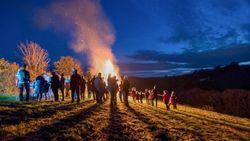 A group of people, including children, gather on a grassy hill at night to watch a large bonfire, with sparks rising into the dark blue sky.