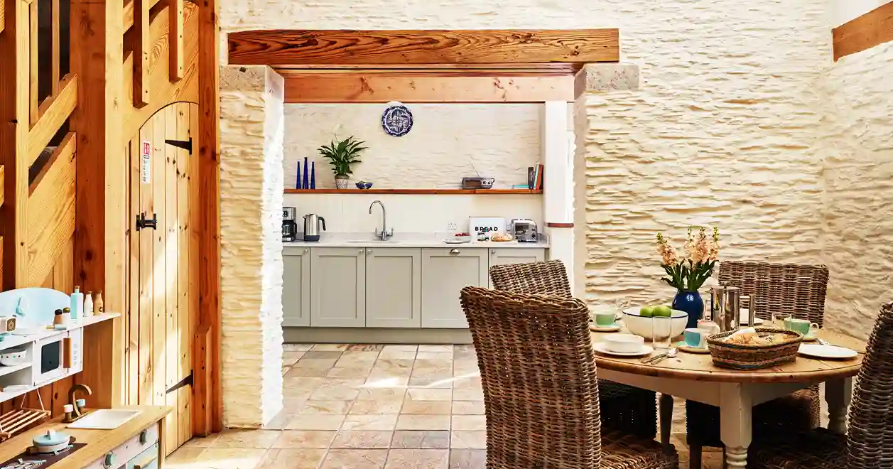 View of The Linhay kitchen at Flear Farm Cottages, featuring wicker dining chairs, pale cabinets, and rustic wood beams.