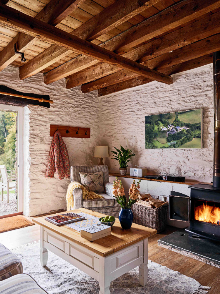 Lounge in 1843 at Flear Farm Cottages with exposed beams, wood burner, armchair, and a coffee table with fresh flowers near open doors to the garden.