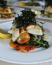 A plated dish of grilled white fish on a bed of greens and roasted potatoes, topped with crispy seaweed, served with a lemon wedge and a drizzle of sauce.