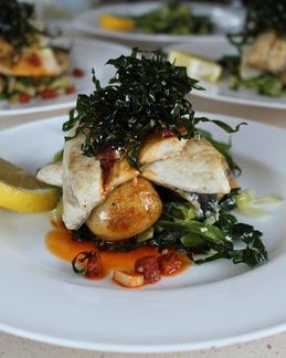 A plated dish of grilled white fish on a bed of greens and roasted potatoes, topped with crispy seaweed, served with a lemon wedge and a drizzle of sauce.