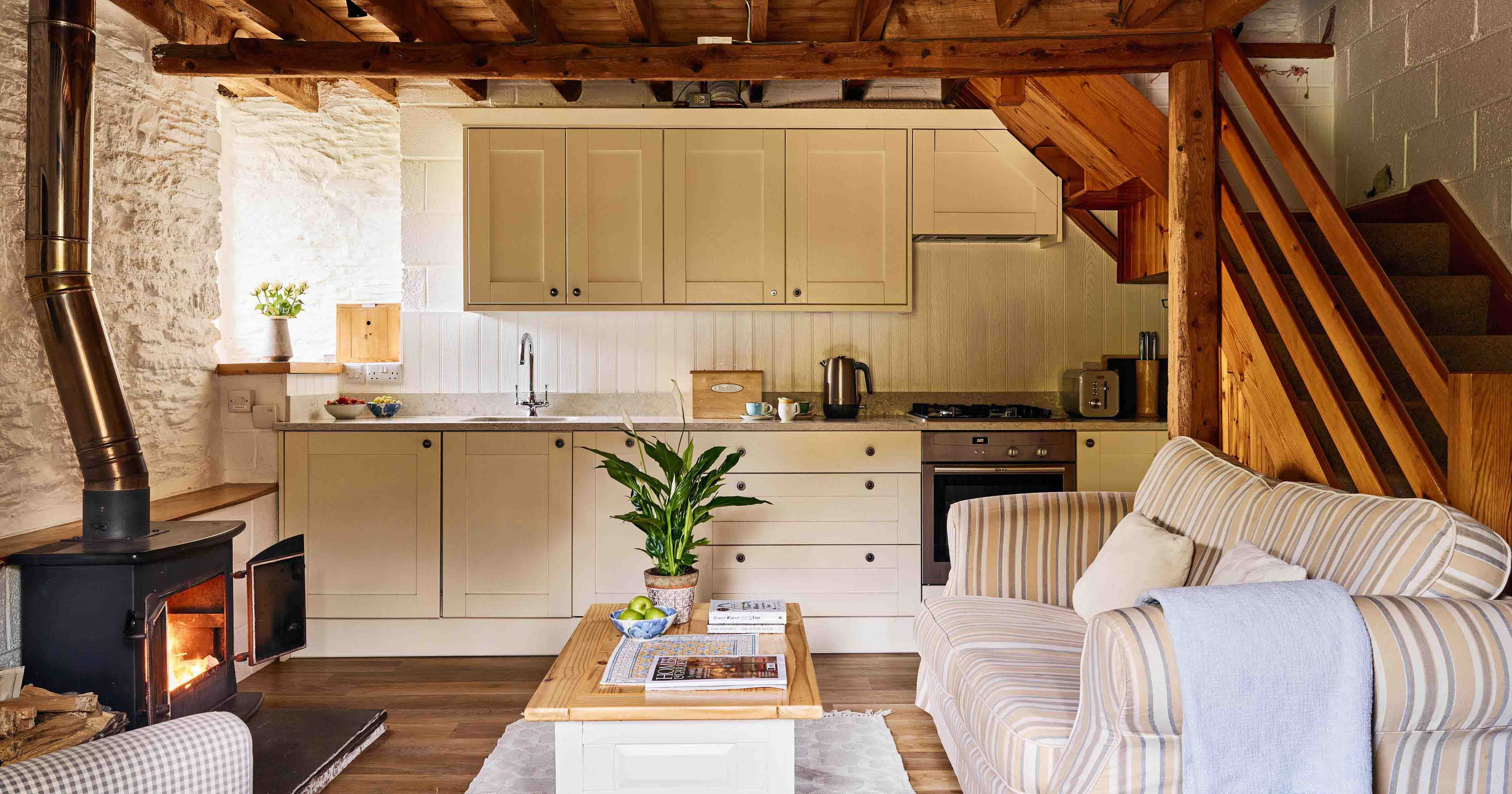 Open-plan kitchen and lounge in 1843 at Flear Farm Cottages, with wood burner, soft seating, striped sofa, and light cabinetry under exposed beams.