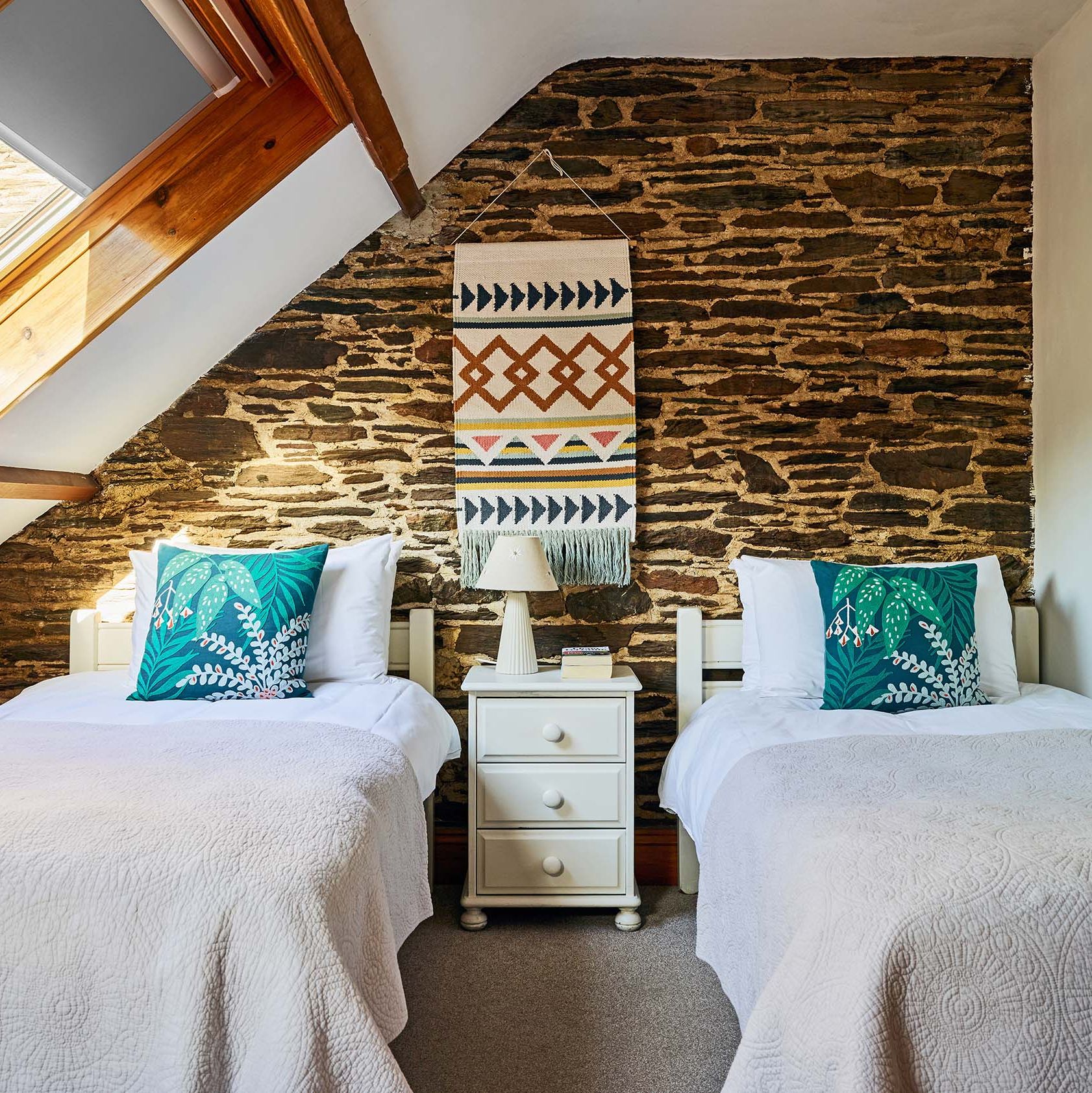 Twin bedroom in Cartwheel at Flear Farm Cottages, with exposed stone wall, skylight, white bedding, and bold green cushions for a fresh touch.