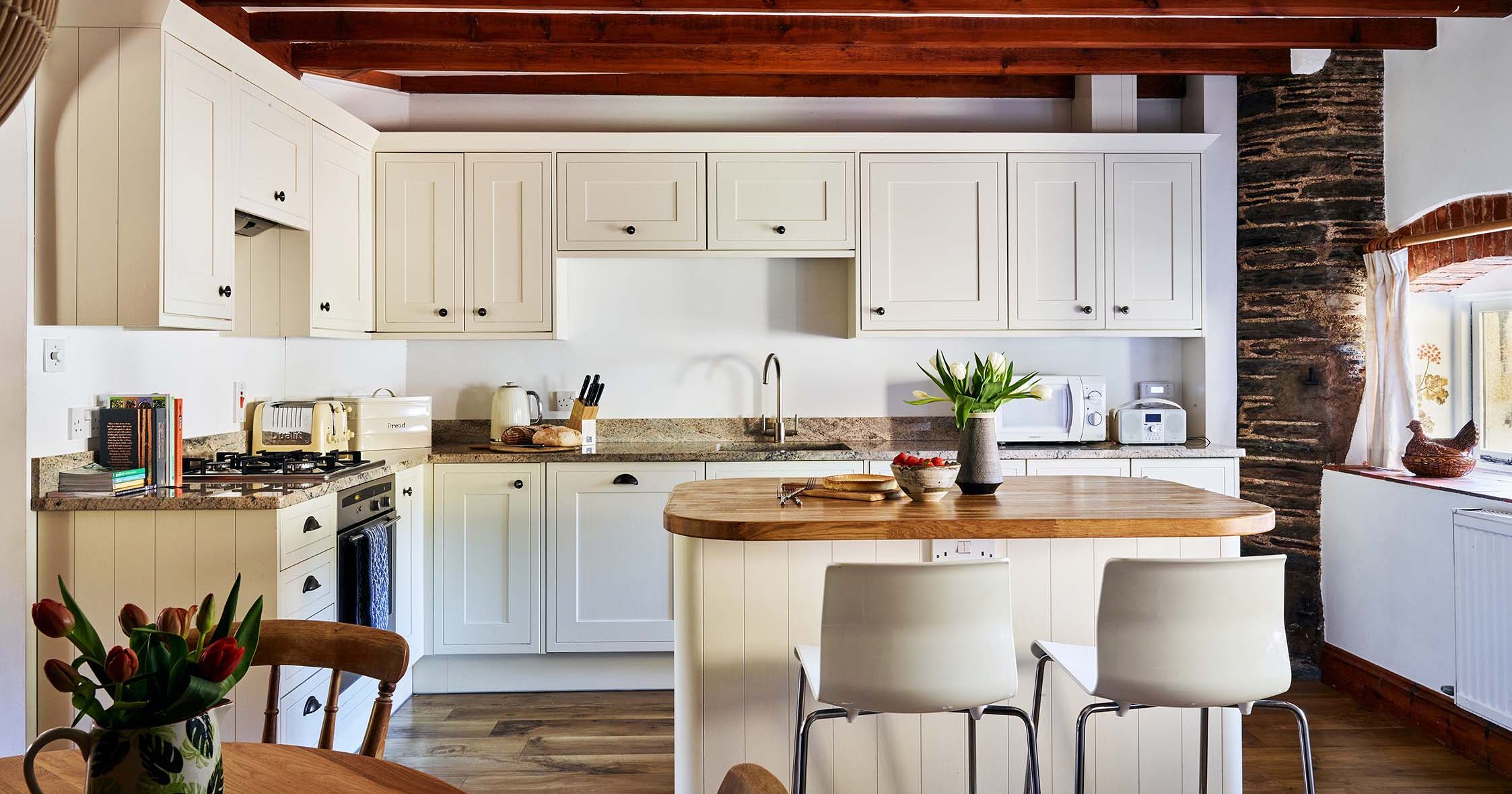 Spacious kitchen in Cartwheel at Flear Farm Cottages, with cream cabinets, central island, exposed beams, and a cosy dining table with fresh flowers.