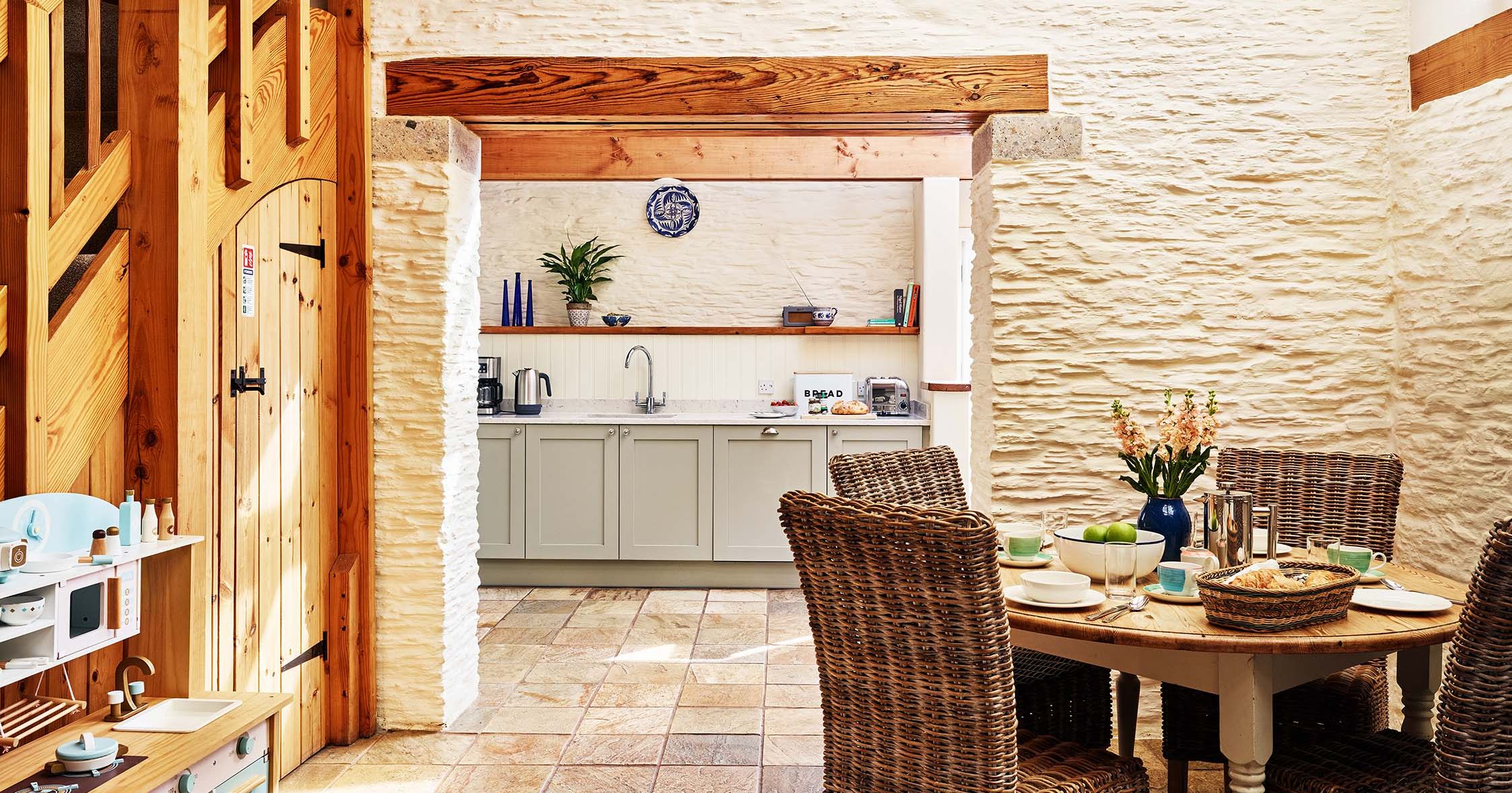 View of The Linhay kitchen at Flear Farm Cottages, featuring wicker dining chairs, pale cabinets, and rustic wood beams.