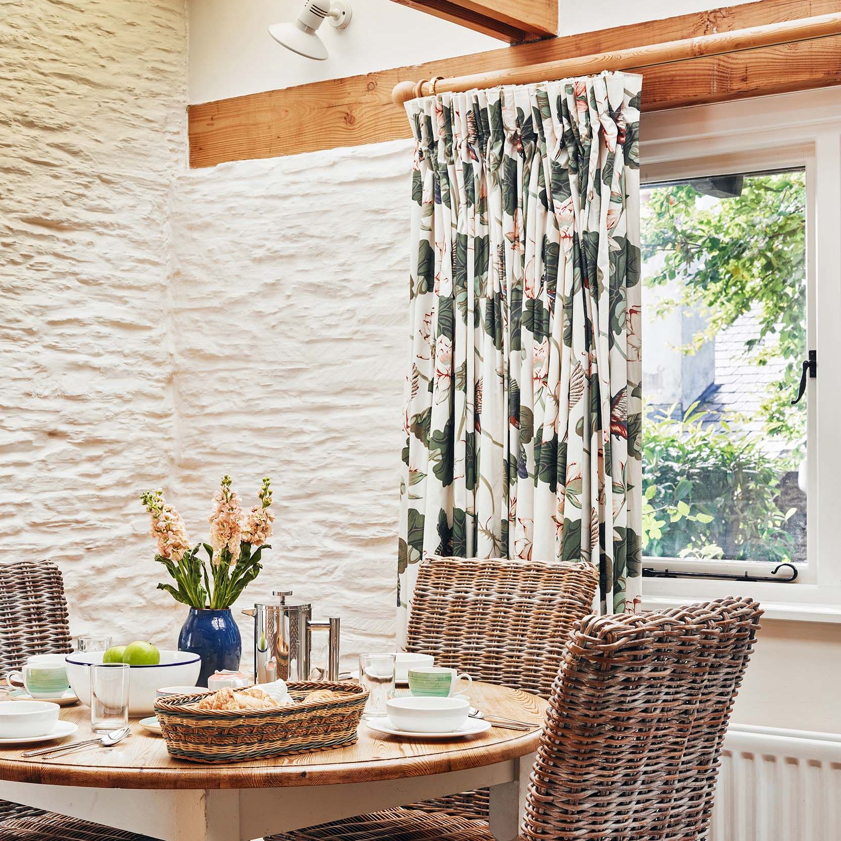 Portrait view of The Linhay dining area at Flear Farm Cottages with wicker chairs, floral curtains, and a set table by the window.
