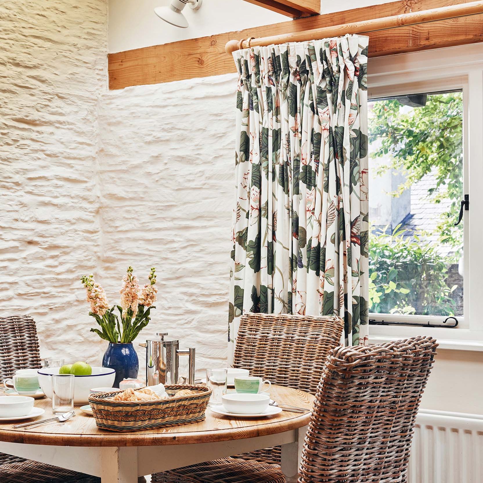 Portrait view of The Linhay dining area at Flear Farm Cottages with wicker chairs, floral curtains, and a set table by the window.