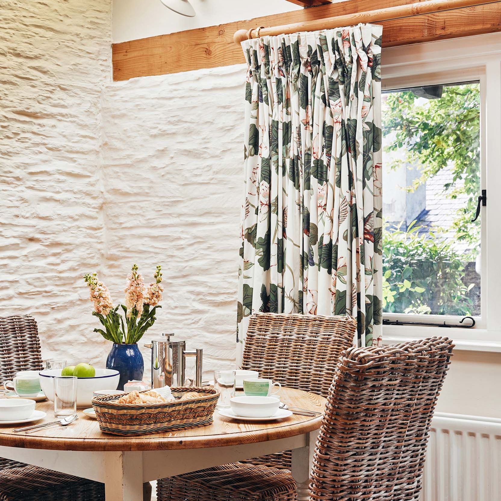 Portrait view of The Linhay dining area at Flear Farm Cottages with wicker chairs, floral curtains, and a set table by the window.