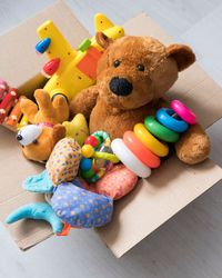 A cardboard box filled with toys, including a teddy bear, stacking rings, and a toy airplane, sits on a light wooden floor with more plush toys and vehicles scattered nearby.
