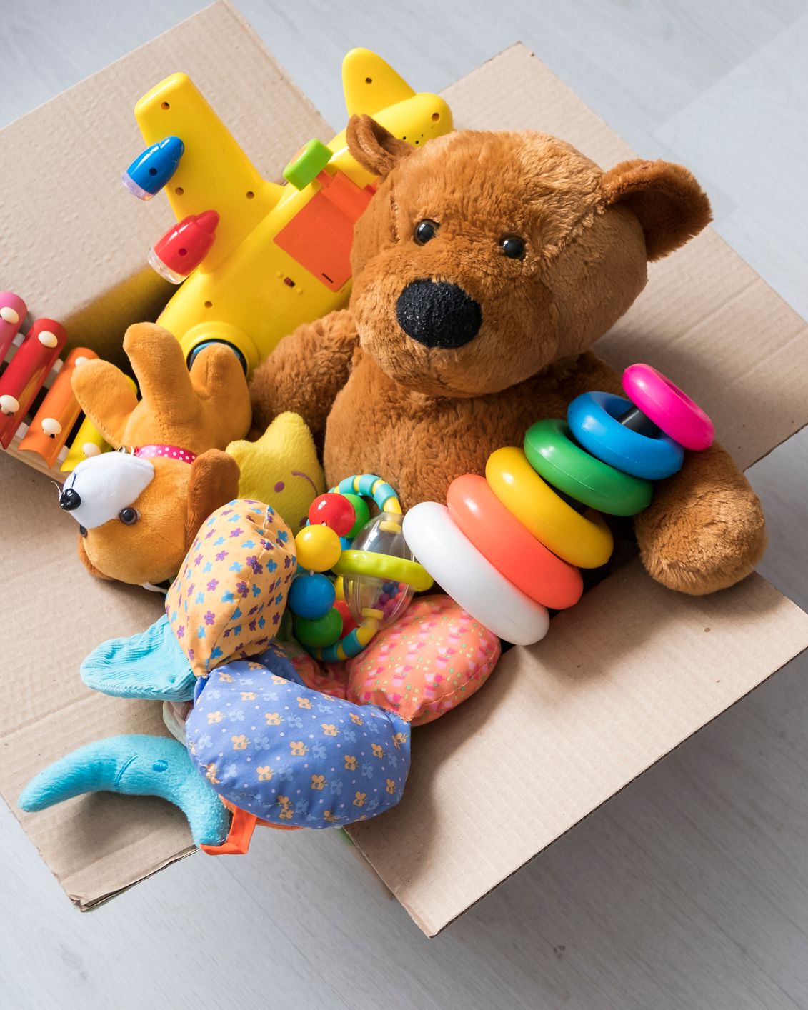 A cardboard box filled with toys, including a teddy bear, stacking rings, and a toy airplane, sits on a light wooden floor with more plush toys and vehicles scattered nearby.