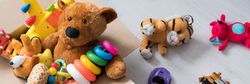 A cardboard box filled with toys, including a teddy bear, stacking rings, and a toy airplane, sits on a light wooden floor with more plush toys and vehicles scattered nearby.