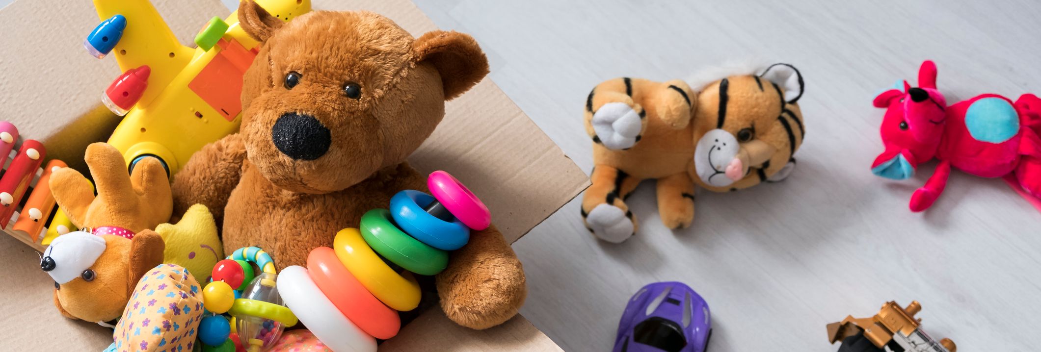 A cardboard box filled with toys, including a teddy bear, stacking rings, and a toy airplane, sits on a light wooden floor with more plush toys and vehicles scattered nearby.