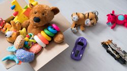 A cardboard box filled with toys, including a teddy bear, stacking rings, and a toy airplane, sits on a light wooden floor with more plush toys and vehicles scattered nearby.