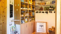 A small farm shop with wooden shelves stocked with local goods, including jams, tea, pasta, and wine, with handwritten labels and a sign for handmade ready meals.