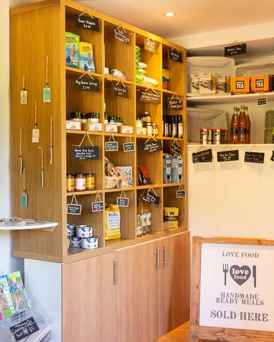 A small farm shop with wooden shelves stocked with local goods, including jams, tea, pasta, and wine, with handwritten labels and a sign for handmade ready meals.