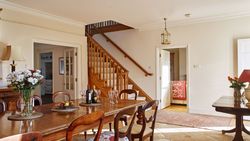 A warm, elegant dining room with a wooden table set with wine, glasses, and flowers, featuring classic chairs, a staircase, and an open doorway leading to a bedroom.