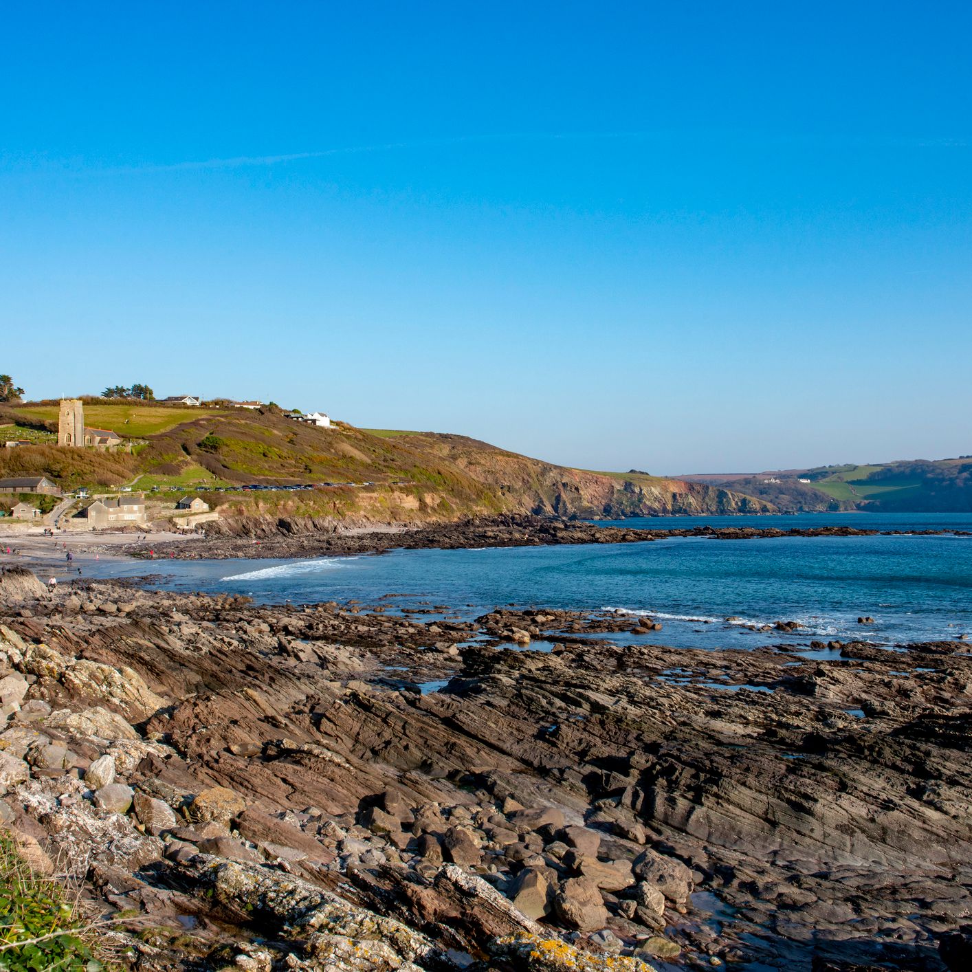 Rocky shoreline at Wembury Beach in Wembury, Devon, with calm blue sea, distant cliffs, and a church tower overlooking the coastal landscape.