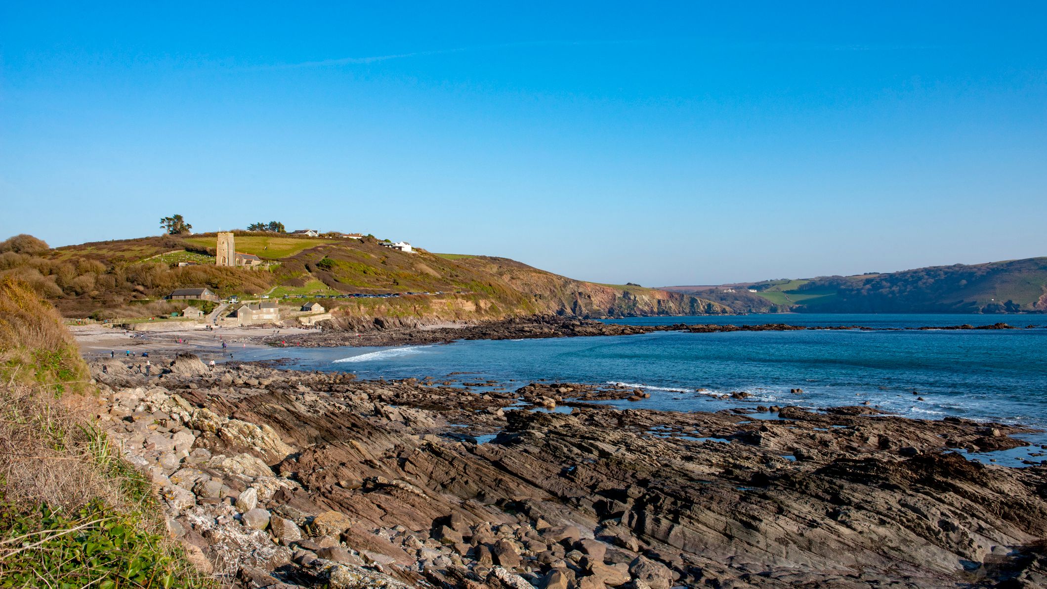 Rocky shoreline at Wembury Beach in Wembury, Devon, with calm blue sea, distant cliffs, and a church tower overlooking the coastal landscape.