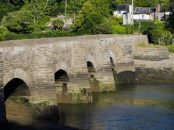 A historic stone bridge with multiple arches spans a calm river, with moss-covered foundations and charming houses nestled among lush greenery in the background.