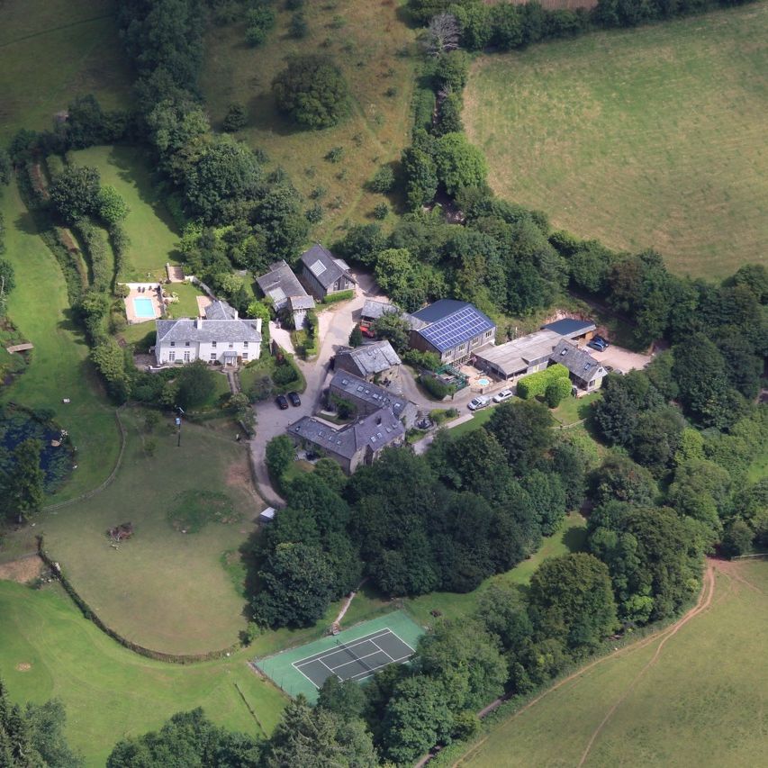 An aerial view of a countryside estate surrounded by lush greenery, featuring multiple buildings, a swimming pool, a tennis court, and open fields nestled among trees.