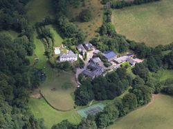 An aerial view of a countryside estate surrounded by lush greenery, featuring multiple buildings, a swimming pool, a tennis court, and open fields nestled among trees.