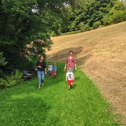 A group of two adults and three children walk along a grassy path beside a small stream, surrounded by lush greenery and a sloping, freshly cut field.